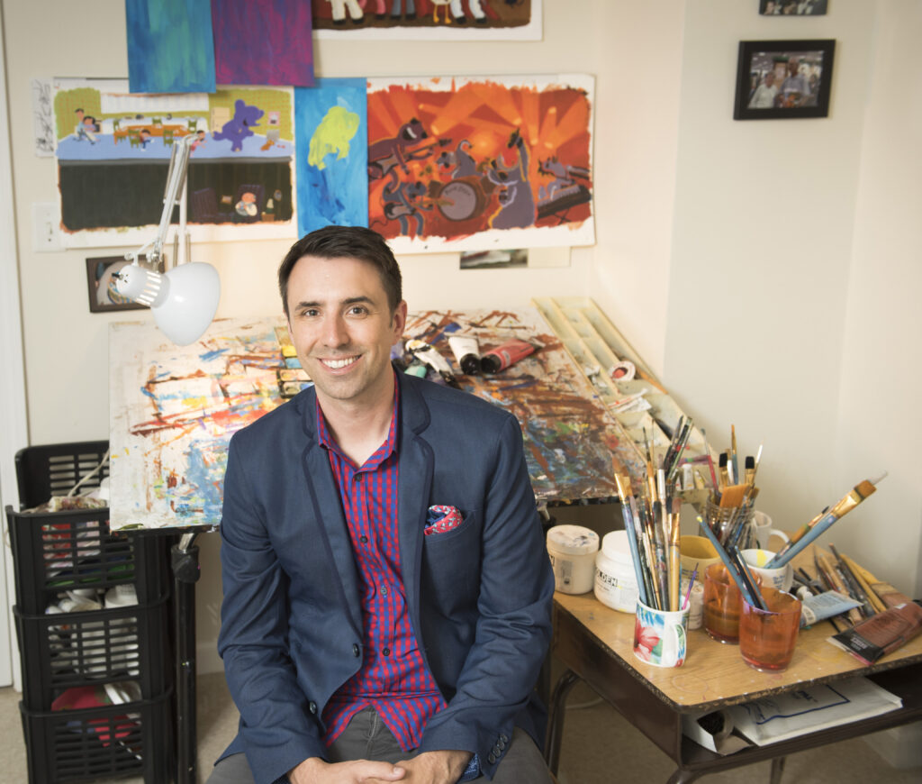 Author Illustrator Jarrett Krosoczka sits in front of his work table covered in paintings. Mugs full of paintbrushes sit next to him and his illustrations are hung on the wall behind him.